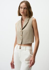 Beige ärmellose cropped Weste mit strukturierter Oberfläche, Einschlagkragen und drei Knöpfen vorne, kombiniert mit weißen hoch geschnittenen Hosen.