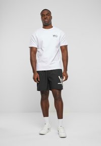 Hvid t-shirt med korte ærmer og trykt logo, parret med sorte shorts med et let abstrakt mønster. Båret med hvide sneakers.