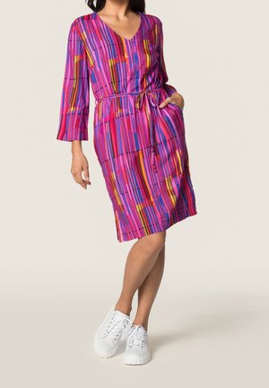 Kvinde iført knælængde, flerfarvet stribet kjole med 3/4-ærmer, V-hals, bindebånd i taljen og hvide sneakers, stående med den ene hånd i lommen.