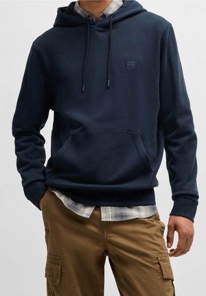 Mężczyzna ubrany w granatową bluzę z kapturem i kieszenią z przodu, nałożoną na koszulę w kratę oraz brązowe spodnie typu cargo, stoi z rękami blisko kieszeni.