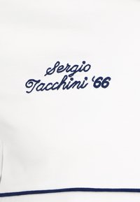 Balts audums ar gludu tekstūru, uz kura ir tumši zilā krāsā izšūts teksts "Sergio Tacchini '66" un horizontāls tumši zils apmales akcentu.