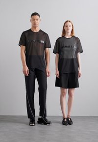 Must must värvilised võrkkangast T-särgid, millel on "HELMUT LANG" tekst, kombineeritud mustade pükstega, millel on valged küljeread, ning must seelik seotud kingadega.