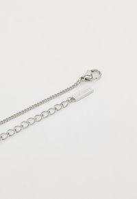 Cadena de metal plateado con un cierre de langosta, que presenta una etiqueta rectangular plana con "LACOSTE" grabada, con formas y texturas de eslabones alternos.