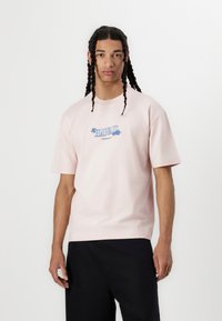 Roza bombažna majica s kratkimi rokavi in okroglim izrezom, ki v središču prikazuje modro grafiko z okrasnimi akcenti.