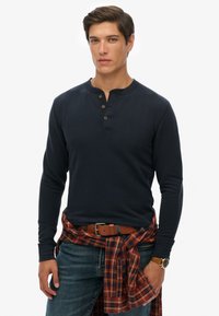 Camisa henley de manga larga en color navy con tapeta de tres botones, combinada con una camisa de cuadros atada a la cintura y jeans de denim azul. Cinturón y reloj marrones.