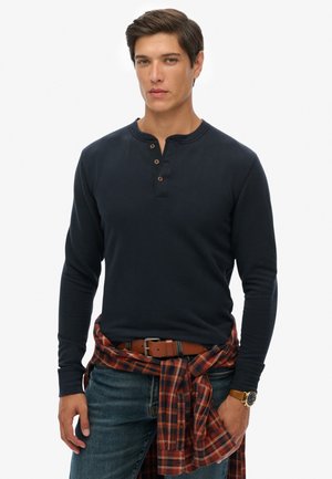 Navy long-sleeve henley shirt with three-button placket, paired with a plaid shirt tied around the waist and blue denim jeans. Brown belt and watch.