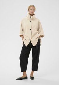 Beige oversized frakke med høj krave og sorte knapper, parret med sorte tapered bukser og flade sorte sko. Glat tekstur, moderne design.