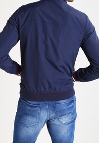 Man som vänder sig bort iförd en marinblå jacka och blå, blekta jeans med händerna vilande på höfterna mot en enfärgad bakgrund.