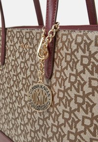 Gros plan sur un sac cabas DKNY monogramme marron avec des poignées en cuir bordeaux et un pendentif chaîne en or suspendu gravé de Donna Karan Chicago 1989.