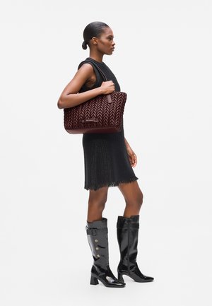 Maroon braided leather tote bag with dual handles, featuring a logo plate and textured pattern. Model wears a black dress and shiny black boots.
