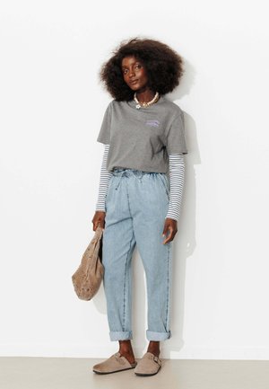 Femme avec une coiffure afro, debout contre un mur blanc, portant un t-shirt gris superposé, des manches longues rayées, un jean bleu clair retroussé, des chaussons beiges et tenant un sac beige.