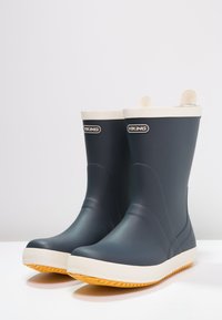 Námornícke modré gumové čižmy s bielym horným pásom a oranžovou textúrovanou podrážkou. Obsahujú logo na hornej prednej časti a ťahacie úchyty pre jednoduché obúvanie.