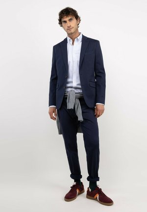 Hombre con traje azul marino, camisa blanca, suéter gris atado alrededor de la cintura, pantalones remangados y zapatillas burdeos, de pie frente a fondo blanco.