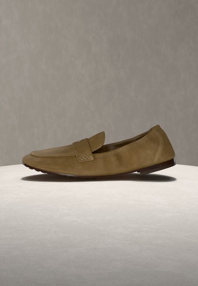 Φωτεινό καφέ σουέντ μοκασίνι με λουρί στο επάνω μέρος, χαμηλό τακούνι και στρογγυλεμένη μύτη, εμφανιζόμενο σε ουδέτερο φόντο και επιφάνεια.