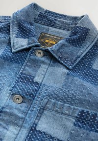 Chemise en denim avec un design en patchwork dans des tons de bleu variés, présentant des motifs texturés, un col pointu et du matériel en métal pour les boutons.