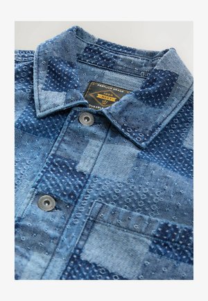 Chemise en denim avec un design en patchwork dans des tons de bleu variés, présentant des motifs texturés, un col pointu et du matériel en métal pour les boutons.