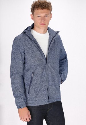 Lichtgewicht blauwe jas met patroon, voorzien van een capuchon, ritszakken en elastische manchetten, gedragen over een witte shirt en donkere jeans.