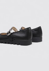 Zapatos de cuero negros con punta redondeada, correa ajustable con hebilla y suela de goma texturizada con un patrón en zigzag.