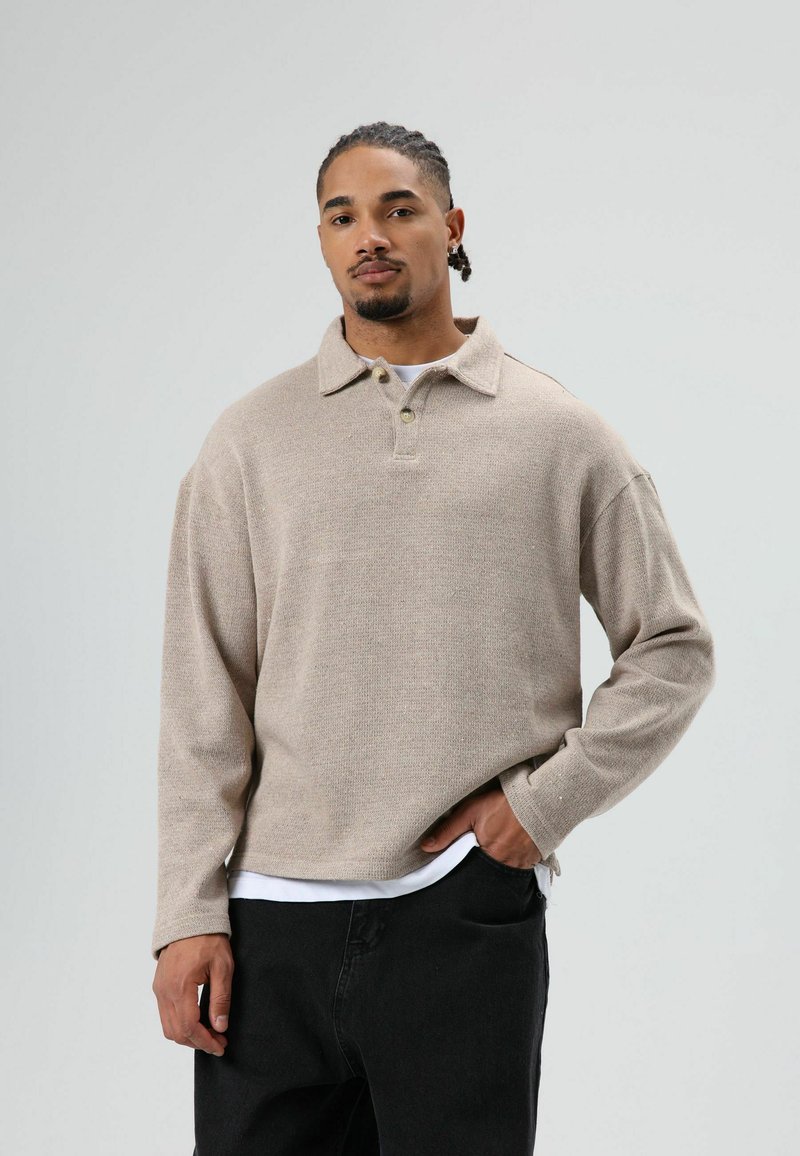 Pull-over en tricot beige avec col, manches longues et coupe décontractée ; comporte deux boutons au niveau du col et une texture subtile. Porté sur un t-shirt blanc.