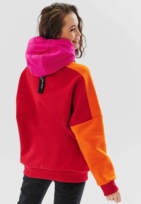 Barevná mikina s červeným tělem, růžovou kapucí a oranžovými manžetami, vyrobená z měkké látky s žebrovaným lemem a manžetami.