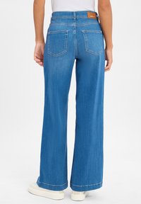 Szerokie niebieskie dżinsy z wysokim stanem, dwoma tylnymi kieszeniami i beżową skórzaną naszywką na pasku. Teksturowany, klasyczny materiał dżinsowy.