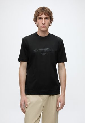 Jeune homme aux cheveux bouclés portant un t-shirt noir avec le contour sombre d'une voiture et un pantalon beige, debout devant un fond blanc uni.