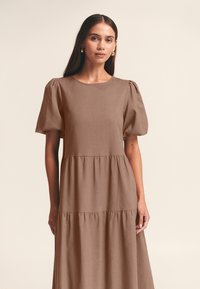 Brun kjole med pufærmer lavet af letvægtsstof, med rund halsudskæring, lagdelt nederdel og blød tekstur. Elegant og enkel design.