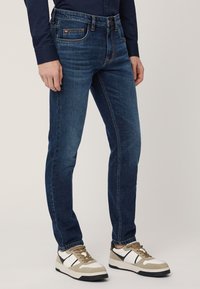 Jeans in denim blu scuro con vestibilità slim, caratterizzati da cuciture visibili, cinque tasche e una superficie usurata. Abbinati a sneaker color crema e grigio.