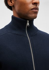 Κοντινό πλάνο ενός ατόμου που φοράει ένα σκούρο ναυτικό μπλουζάκι με ραβδώσεις και φερμουάρ, με το φερμουάρ κλειστό εντελώς μέχρι ψηλά στο γιακά.