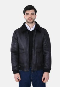 Uomo che indossa una giacca di shearling nera sopra una camicia bianca e un maglione nero, in piedi davanti a uno sfondo chiaro e uniforme.