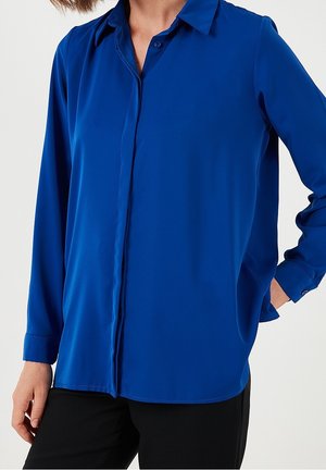 Kobieta w długim rękawie, zapinanej na guziki, królewskoniebieskiej bluzce i czarnych spodniach, stojąca z jedną ręką w kieszeni na jednolitym tle.