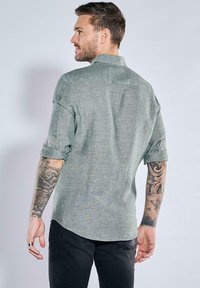 Hellgrüne Hemdbluse aus texturiertem Stoff mit hochgerollten Ärmeln und Kragen, von hinten an einem Modell präsentiert.