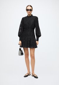 Black lace dress with long puff sleeves, a high neck, and button closure. Features a flared skirt and a ruffled hem. Worn with black ballet flats and a small handbag.