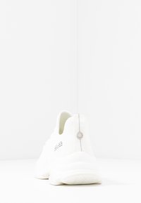 Valkoinen sneaker, jossa on teksturoitu verkko-osa yläosassa, pehmustettu nilkkapanta ja kumipohja. Sisältää metallisen koristeen kantapäässä ja logotiedot sivulla.