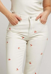 Jeans en denim blanc avec des motifs de cerises rouges brodés. Taille haute avec poches avant et silhouette ajustée.