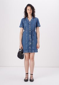 Kratka denim obleka v modri barvi, z gumbnim zapenjanjem spredaj, pasom in žepi. Ujemajoča mala črna pletena torbica.