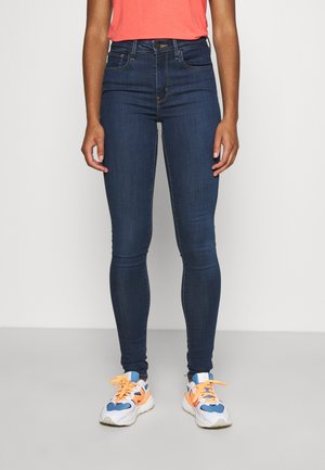 Jeans ajustados de color azul oscuro con una cintura de altura media, cinco bolsillos y detalles de costura sutiles, combinados con zapatillas blancas y naranjas.