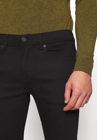 Svarta jeans med en mjuk textur och klassisk passform, med knappstängning och femficksdesign, kombinerade med en grön stickad tröja.