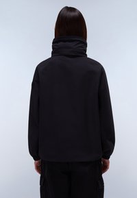 Persona con cabello liso y oscuro, que lleva una chaqueta negra de cuello alto y pantalones cargo negros, mostrada de espaldas contra un fondo liso.