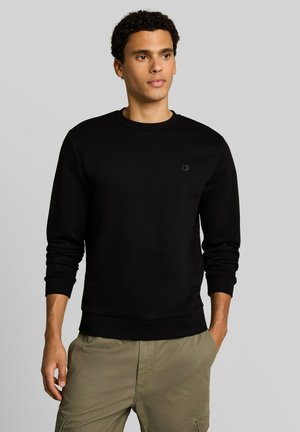 Jonge man met kort krullend haar, gekleed in een zwart sweatshirt met lange mouwen en kaki broek, staand tegen een simpele lichte achtergrond.