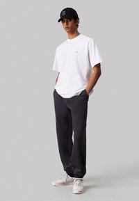 T-shirt bianco a maniche corte con un piccolo logo, abbinato a pantaloni tuta grigio scuro e scarpe da ginnastica beige. Cappellino nero con dettagli testuali.