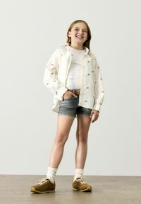 Camisa blanca de botones con motivos en azul marino, combinada con pantalones cortos de mezclilla gris claro. Zapatillas marrones y calcetines blancos completan el conjunto.