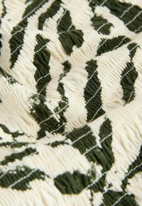 Tissu texturé avec une base crème ornée de motifs de feuilles vert foncé, design froncé et surpiqûres blanches contrastantes sur toute la surface.