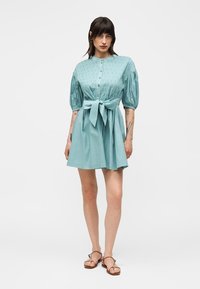 Ženska stoji in nosi svetlo modro kratko obleko z napihnjenimi rokavi, gumbi spredaj, pasom v pasu in rjavimi ravnimi sandali.