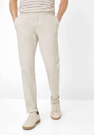 Uomo che indossa pantaloni chino beige con risvolti, sneakers beige e una camicia bianca a maniche corte con sottili righe rosse infilata dentro i pantaloni.