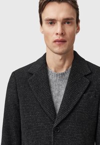 Svart texturerad kappa med spetsig krage och enkel knappstängning, lager-på-lager över en ljusgrå, stickad tröja. Enkel, modern design.