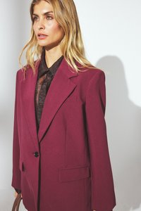 Blazer oversized de couleur vin avec des revers notés, fermeture à un bouton et poches latérales. Tissu texturé, associé à une chemise noire.