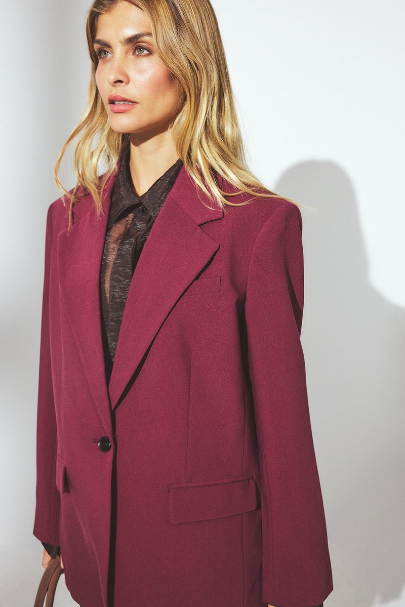Blazer oversized de couleur vin avec des revers notés, fermeture à un bouton et poches latérales. Tissu texturé, associé à une chemise noire.