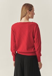 Piros kötött pulóver gyémántmintával és bordázott szegéllyel. Esés vállakkal és széles nyakkivágással rendelkezik. Fekete nadrággal van párosítva.