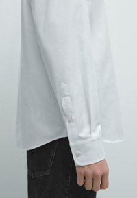 Camicia bianca a maniche lunghe con bottoni, realizzata in tessuto liscio. Presenta polsini con due bottoni e un dettaglio di taglio sottile al polso.